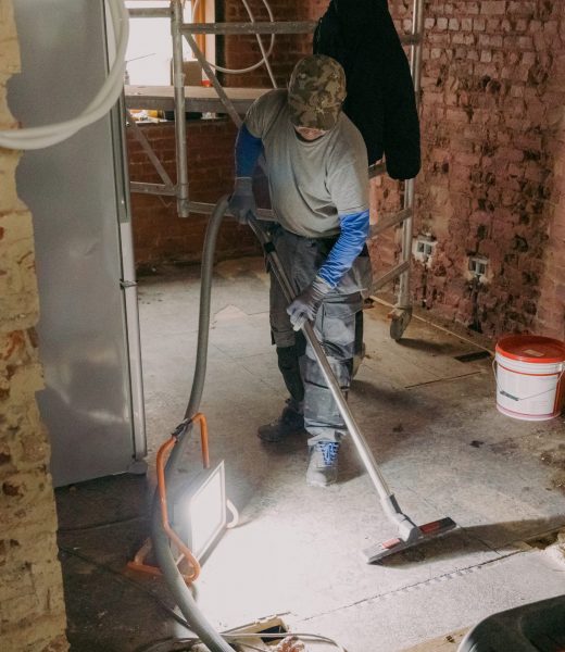 A young man vacuums the landing in a repair room. One young Caucasian man in a gray casual work uniform vacuums up construction debris after laying bricks in a room where renovations are underway, standing under the light of a lantern in an old ruined house, close-up side view.