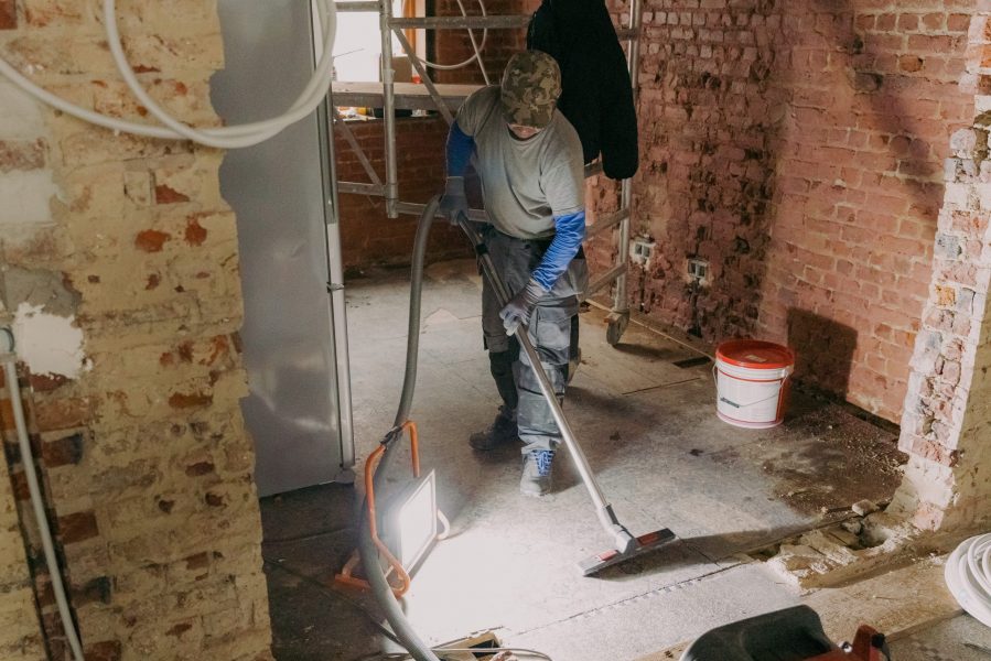 A young man vacuums the landing in a repair room. One young Caucasian man in a gray casual work uniform vacuums up construction debris after laying bricks in a room where renovations are underway, standing under the light of a lantern in an old ruined house, close-up side view.